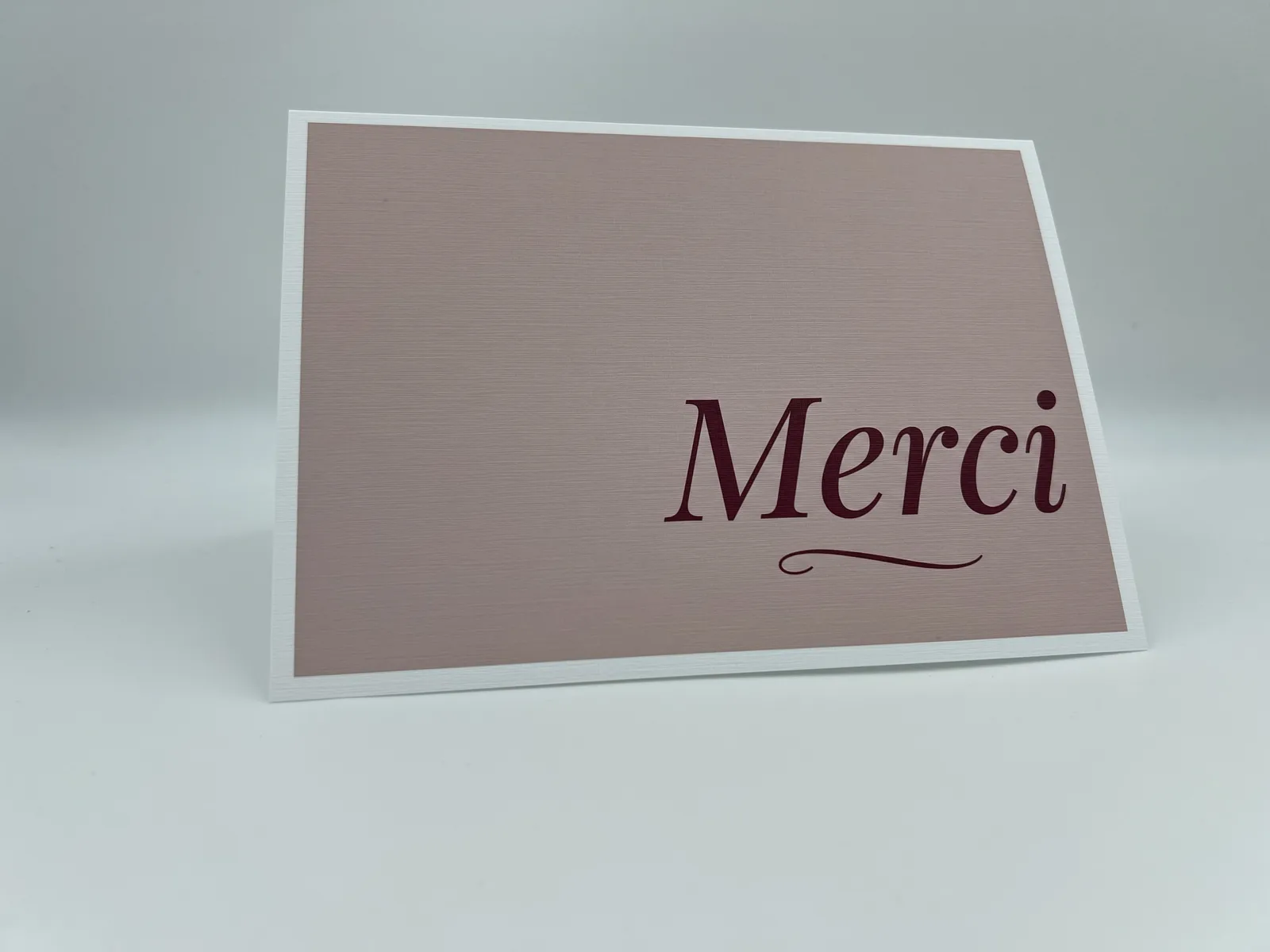 A folded thank-you card reading Merci in burgundy script on a dusty pink panel