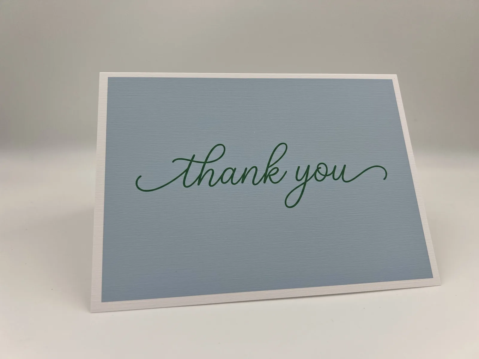 A handwritten thank-you card with "thank you" lettered in green script on a dusty-blue background