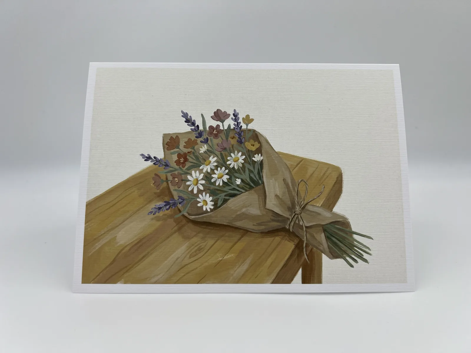 A wedding thank-you card painted with a wildflower bouquet wrapped in kraft paper on a wooden bench